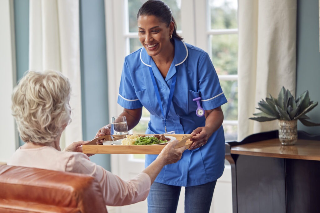 female care worker in uniform bringing meal on tray to senior woman sitting in lounge at home